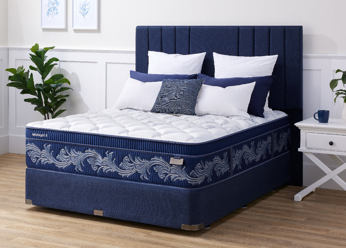 A Sleepyhead mattress set in a navy blue upholstered bed frame with a matching headboard, styled in a bedroom setting with white pillows, a white nightstand, and a green plant.