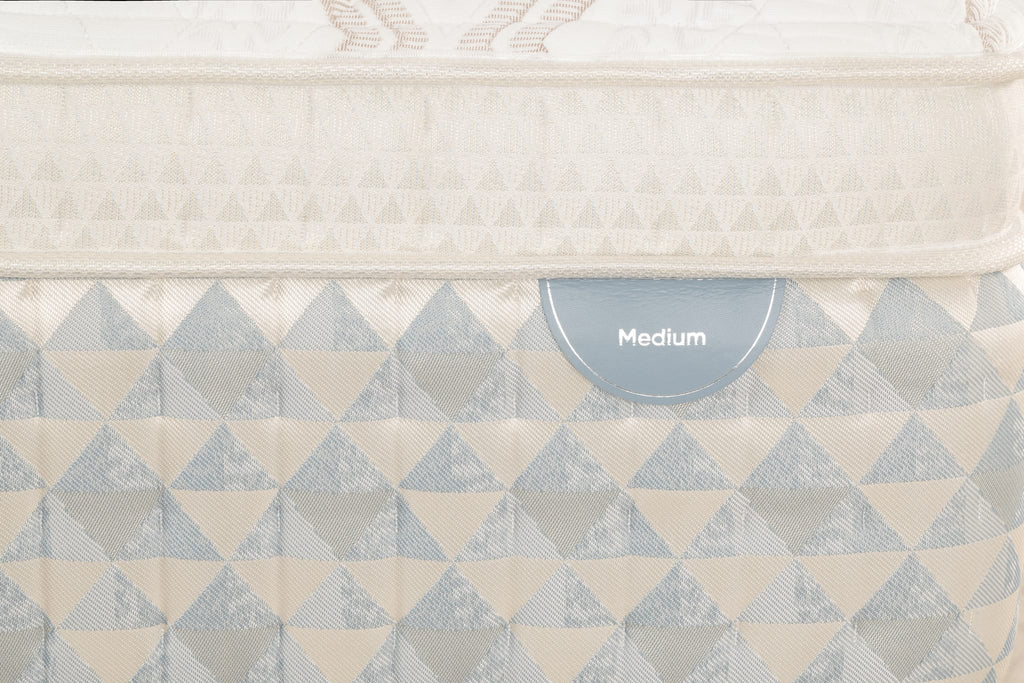 Close-up of a Sleepyhead mattress with a geometric triangle pattern in cream and light blue-gray, featuring a small blue-gray label that reads 'Medium'.