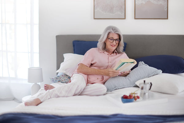An older woman with gray hair and glasses sits comfortably on a bed with white linens and blue pillows, holding and reading a book. The bedroom is bright and airy with a gray headboard, two framed wall art prints, a bedside lamp, and a small tray of fruit and coffee on the bed.