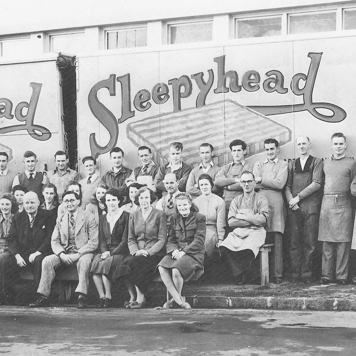 A black and white group photograph of workers posing in front of a large truck with the word 'Sleepyhead' painted across it. Multiple men and women are seated and standing in formal attire from the mid-20th century.