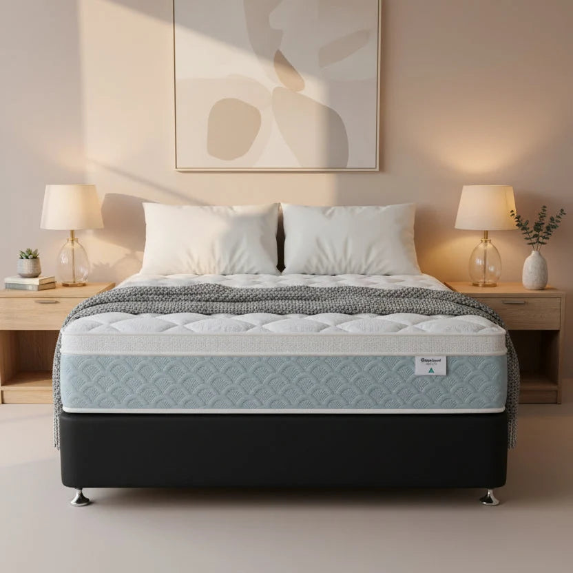 A mattress on a black bed frame in a styled bedroom setting, with white pillows, a gray throw blanket, wooden bedside tables, lamps, and a beige wall with abstract wall art.