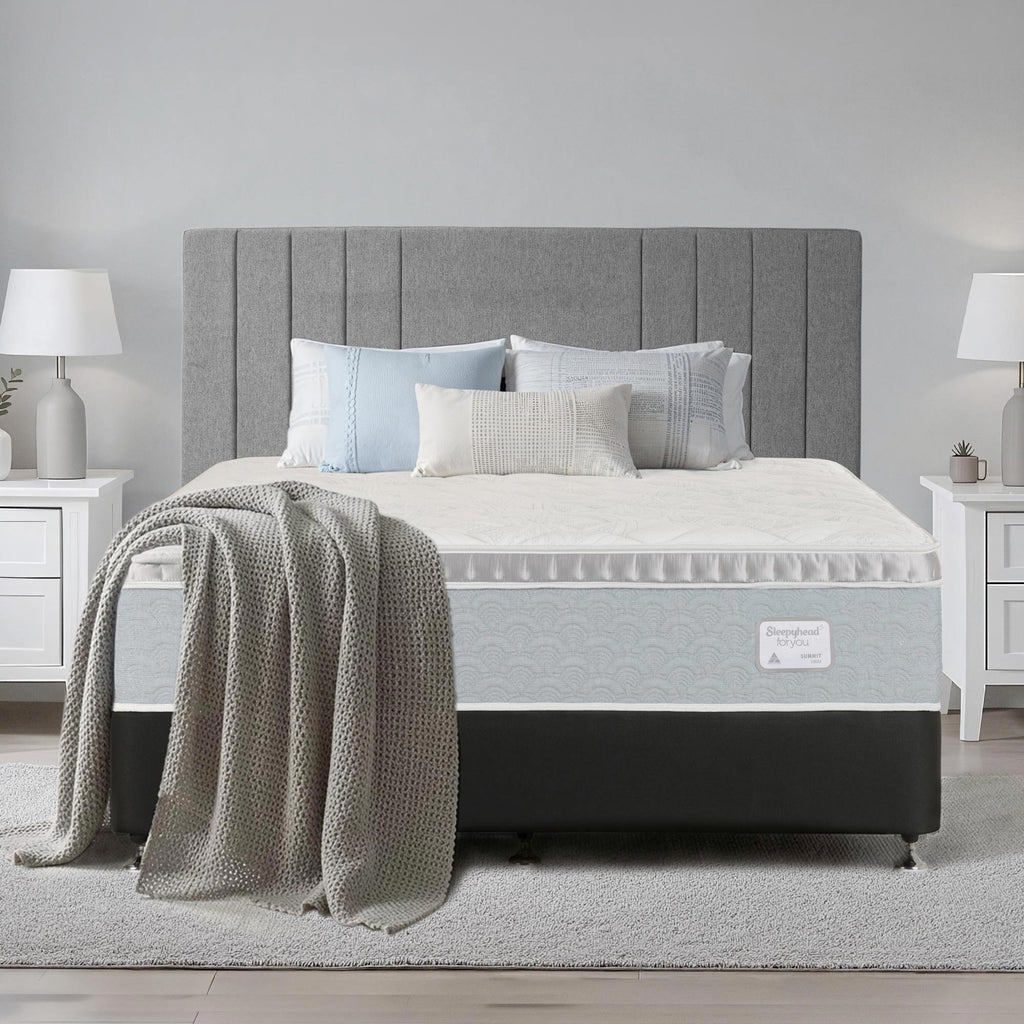 A bed with a gray upholstered headboard, Sleepyhead mattress, black base, and light gray bedding, styled with pillows and a textured throw blanket in a bright bedroom setting.