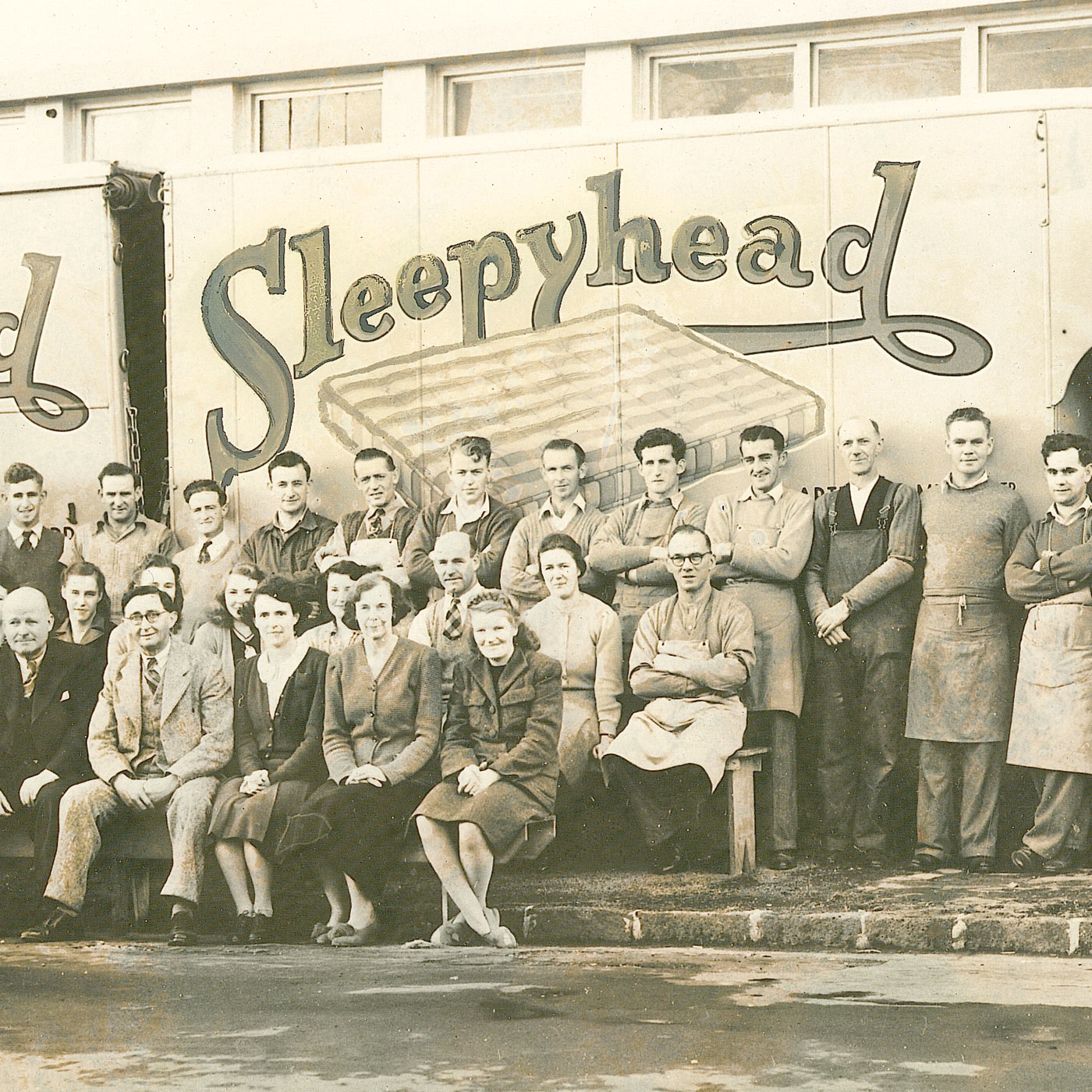 A vintage black-and-white group photograph showing a large team of workers posing in front of a truck with the word 'Sleepyhead' painted across the side.