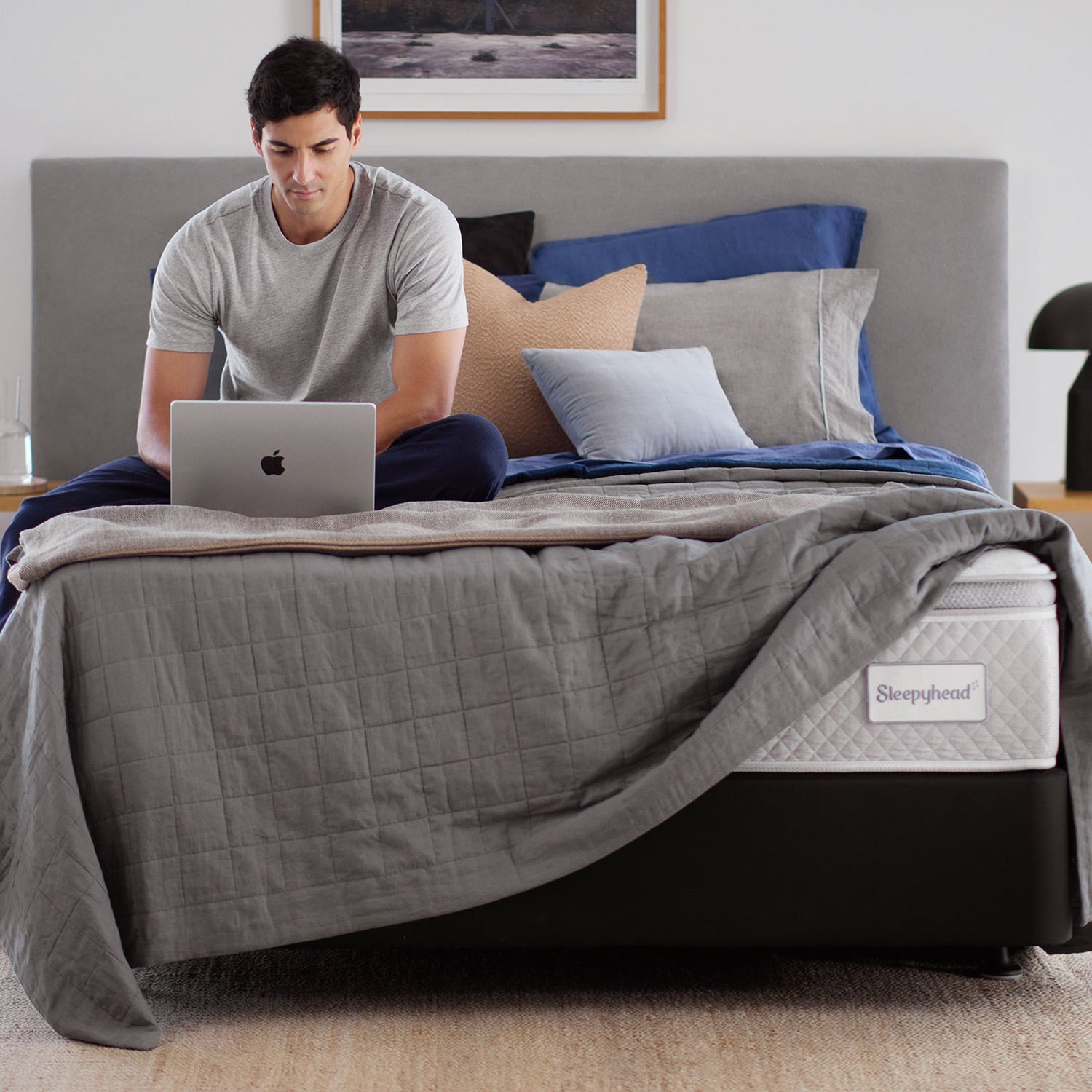 A man sits on a bed using a laptop in a modern bedroom setting. The bed features a gray quilted comforter, blue and gray pillows, and a visible Sleepyhead mattress on a black bed frame.