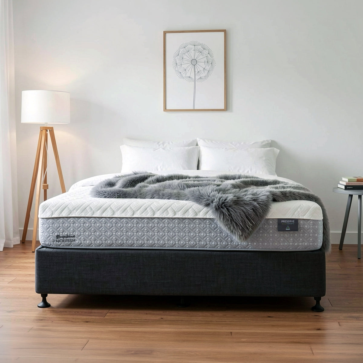 A Sleepyhead mattress on a dark upholstered bed frame in a styled bedroom setting, with white pillows, a gray faux-fur throw blanket, a floor lamp, and a small side table against a white wall.