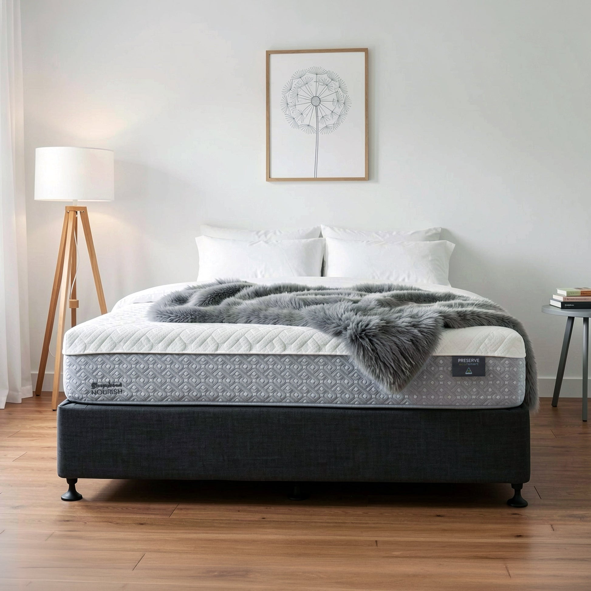 A Sleepyhead mattress on a dark upholstered bed frame in a styled bedroom setting, with white pillows, a gray faux-fur throw blanket, a floor lamp, and a small side table against a white wall.