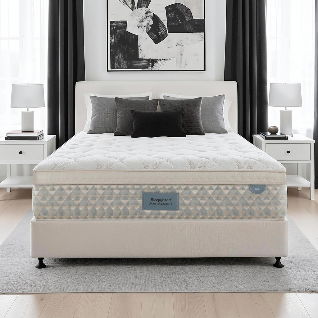 A Sleepyhead mattress on a matching upholstered bed frame, styled in a modern bedroom setting with white nightstands, lamps, gray curtains, and a black and white abstract wall art piece.