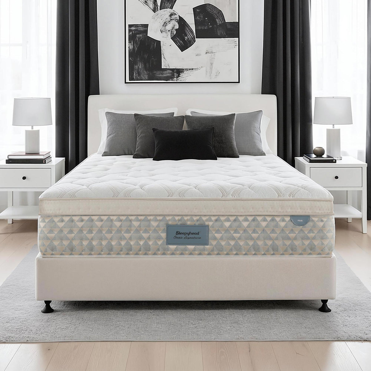 A Sleepyhead mattress on a matching upholstered bed frame, styled in a modern bedroom setting with white nightstands, lamps, gray curtains, and a black and white abstract wall art piece.