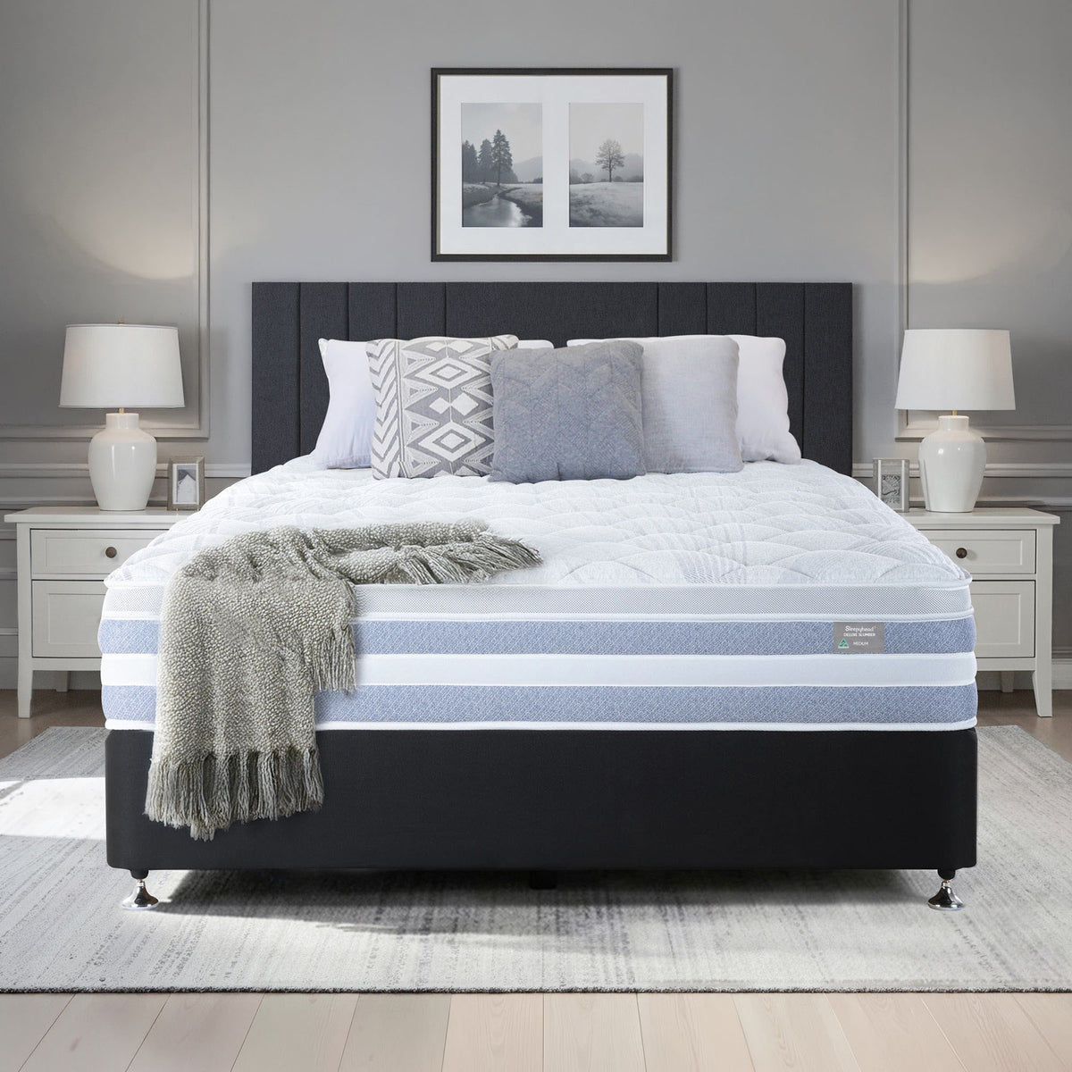 A Sleepyhead bed with a dark upholstered headboard and base, dressed in white bedding with light blue stripes, patterned pillows, a gray throw blanket, and a matching gray rug in a staged bedroom.