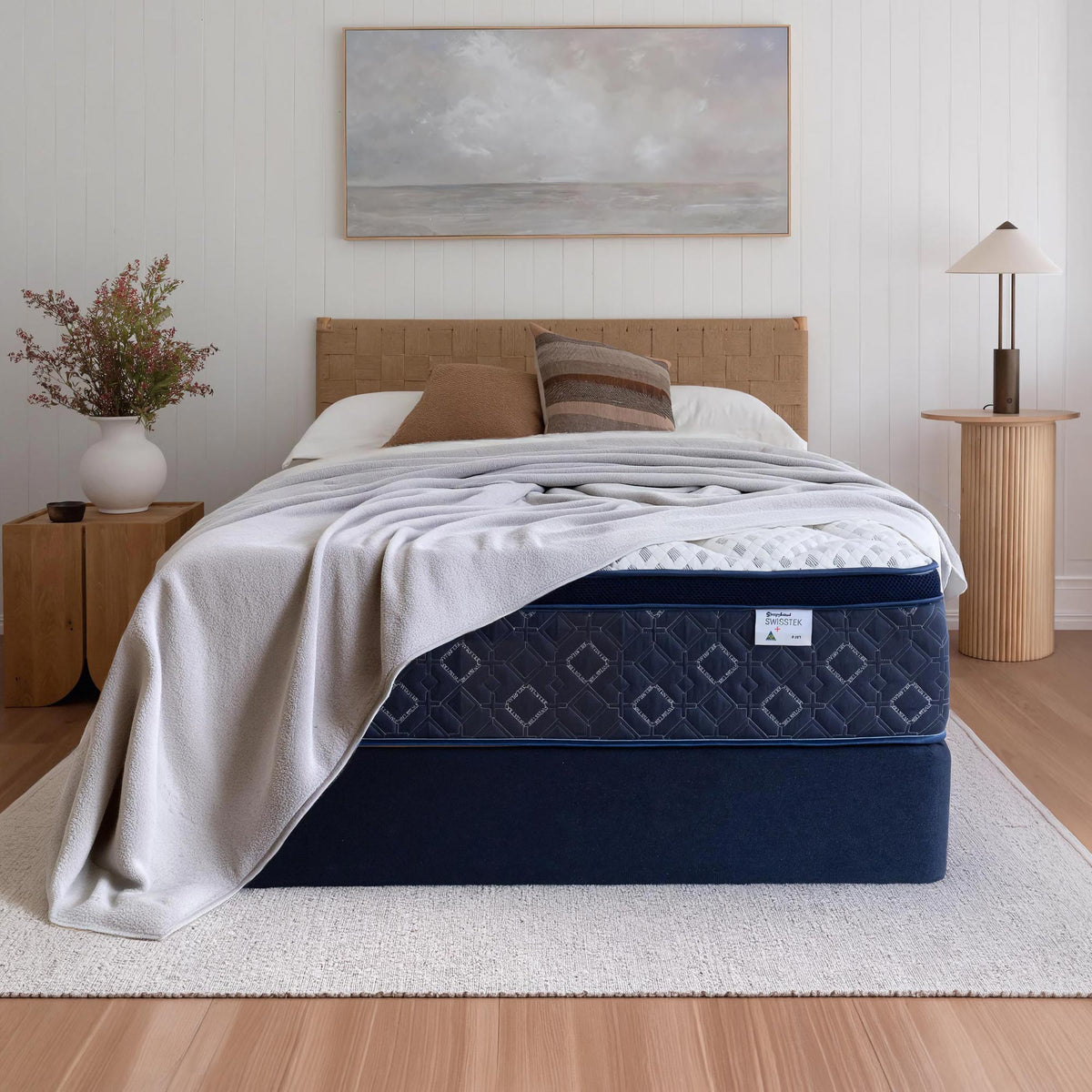 A Sleepyhead bed with a navy blue mattress and box spring, styled with a light gray throw blanket, brown pillows, and a beige headboard in a bright, modern bedroom.
