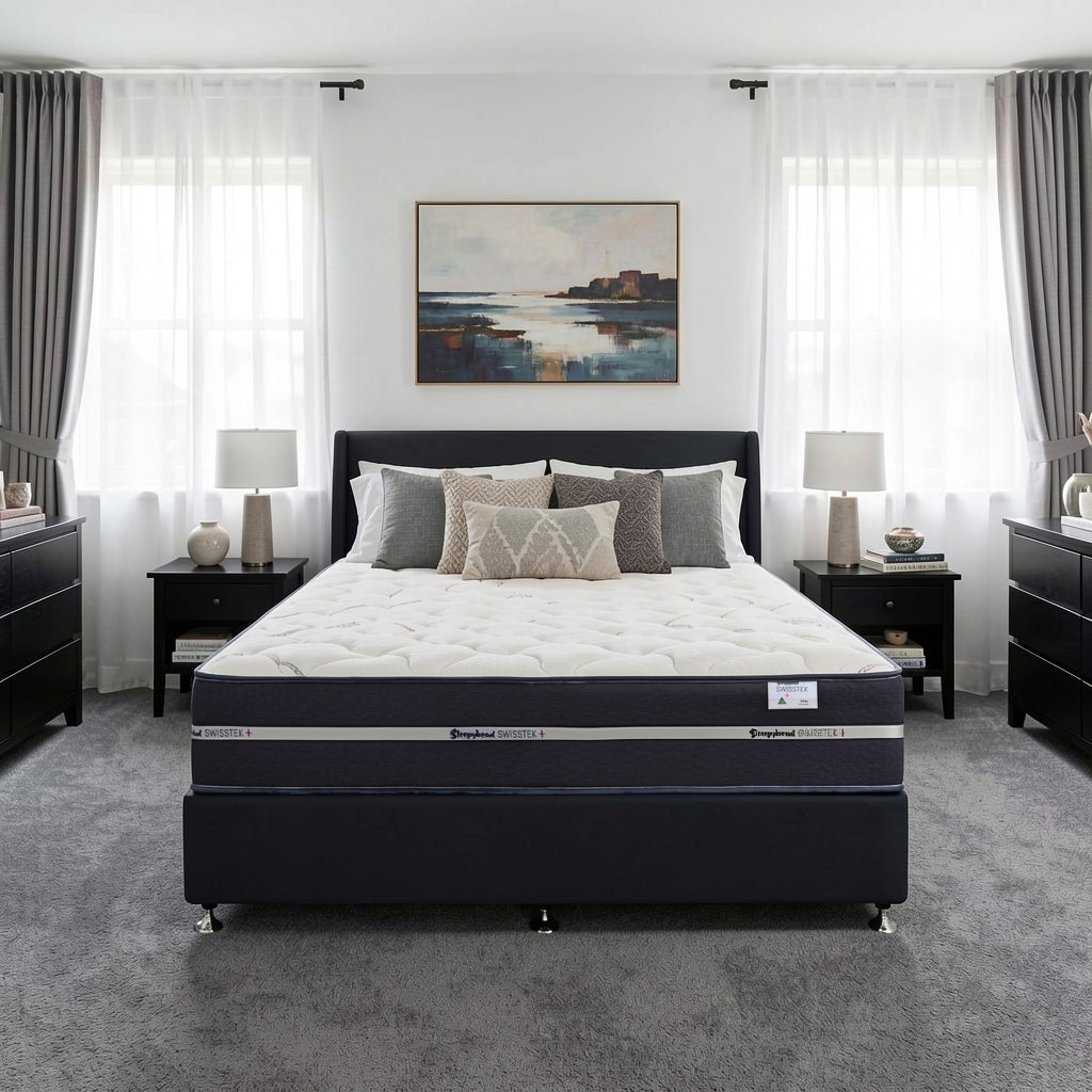 A bed with a dark navy upholstered frame and a Sleepyhead mattress, styled in a modern bedroom with gray curtains, black nightstands, and a landscape painting on the wall.